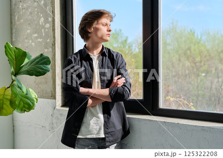 Upset sad teenager guy looking at window with crossed arms Upset sad teenager guy looking at window with crossed arms 125322208