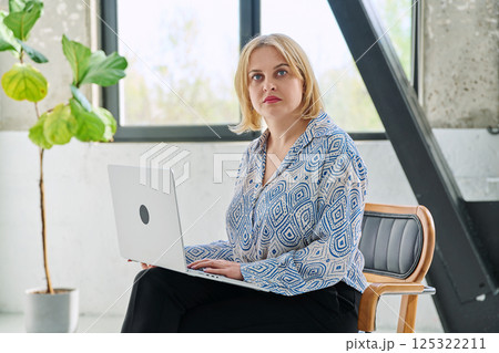 Serious middle aged business woman sitting in chair with laptop looking at camera Serious middle aged business woman sitting in chair with laptop looking at camera 125322211