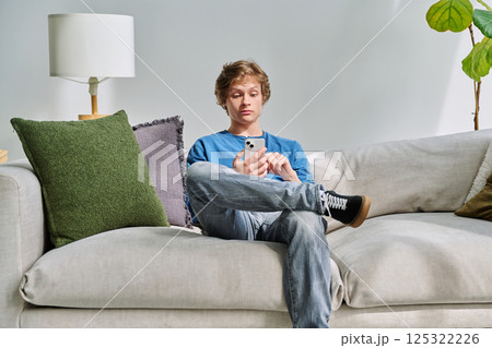 Handsome surprised teenage guy using smartphone sitting on couch at home 125322226