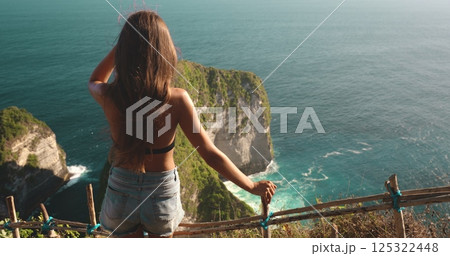 Young woman is taking in the beauty of the turquoise ocean and the dramatic cliffs of kelingking beach from a scenic viewpoint 125322448