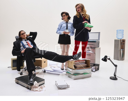 Man leaning back with hands behind head while two women in formal outfits stand holding cup and phone in cluttered workspace. 125323947