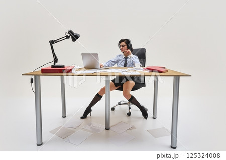Young woman in shirt and tie working with laptop and documents on messy table, wearing headphones and focused expression in office setup. Young woman in shirt and tie working with laptop and documents on messy table, wearing headphones and focused expression in office setup. 125324008