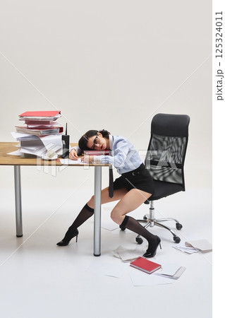 Young woman resting head on documents, leaning on desk with sleepy expression in messy office setup against white studio background. Young woman resting head on documents, leaning on desk with sleepy expression in messy office setup against white studio background. 125324011