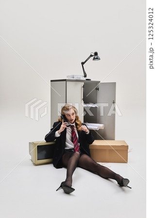 Young woman sitting on floor leaning against cabinet holding phone with exhausted face in messy office scene. 125324012