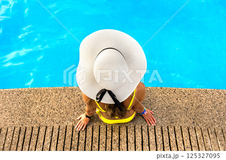 Top down view of young woman wearing yellow straw hat resting near swimming pool with clear blue water on summer sunny day. Top down view of young woman wearing yellow straw hat resting near swimming pool with clear blue water on summer sunny day. 125327095