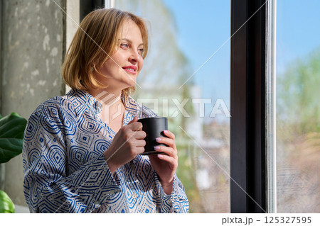 Portrait of beautiful middle-aged woman with cup in her hands looking out window Portrait of beautiful middle-aged woman with cup in her hands looking out window 125327595
