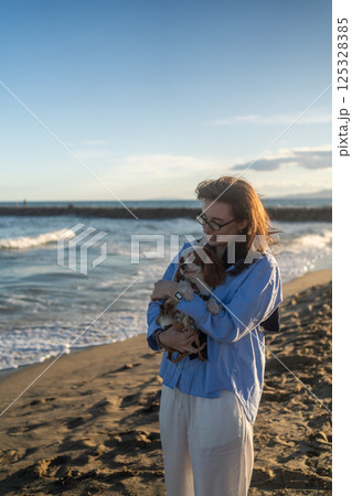 A young woman hugs her Cavalier King Charles Spaniel on the beach at sunset. Warm light, waves, and smiles capture a peaceful, joyful moment of connection and relaxation. 125328385