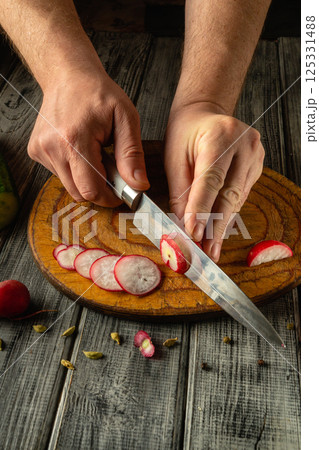 Hands skillfully slice radishes into thin rounds using a sharp knife on a rustic wooden cutting board, highlighting culinary precision and fresh ingredients in a cozy kitchen atmosphere 125331488