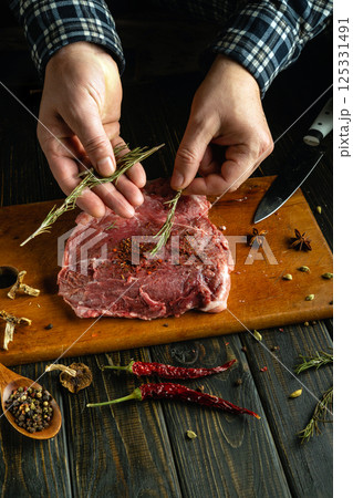Hands carefully sprinkle fresh herbs and spices onto a raw piece of meat on a wooden cutting board. Surrounding ingredients include dried spices and red chili peppers 125331491