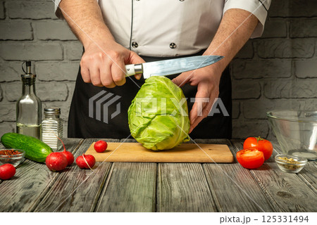 A chef in a white uniform skillfully cuts a head of cabbage on a wooden cutting board surrounded by fresh vegetables like tomatoes, cucumbers, and radishes 125331494