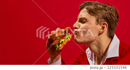 Side view humorous portrait of handsome young man in suit kissing delicious chicken burger against bold red background. Side view humorous portrait of handsome young man in suit kissing delicious chicken burger against bold red background. 125331546