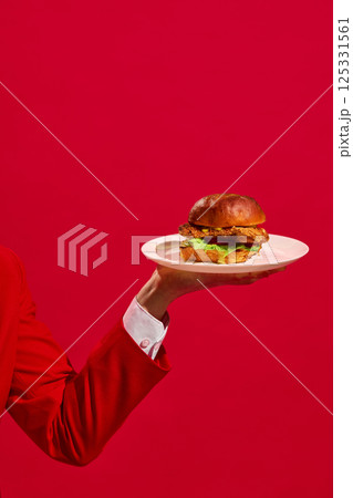 Vertical image of male hand holding white plate with delicious chicken burger against a bright red studio background. Vertical image of male hand holding white plate with delicious chicken burger against a bright red studio background. 125331561