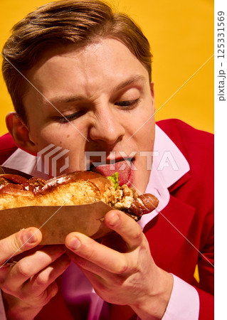 Close-up portrait of young man licking tip of hot dog with exaggerated expression and playful hunger against bright yellow background. 125331569