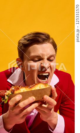 Portrait of young emotional man passionately eating hot dog against vivid yellow background. Close-up. Food passion. 125331570