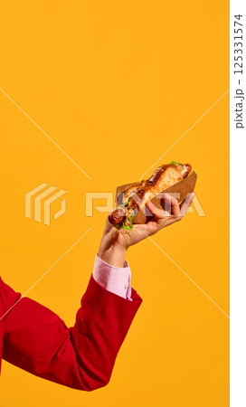 Male hand in red suit holding grilled hot dog against bright yellow background. Vertical image. 125331574