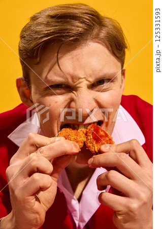 Close-up portrait of handsome young man fiercely biting two chicken pieces with sauce against bold yellow background. Close-up portrait of handsome young man fiercely biting two chicken pieces with sauce against bold yellow background. 125331593