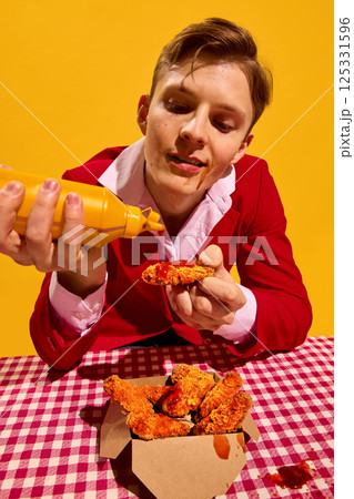 Man squeezing sauce on fried chicken nuggets with humorous expression over checkered tablecloth and bright yellow background. 125331596