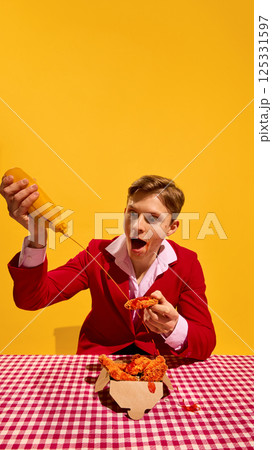 Man holding crispy chicken while pouring mustard sauce with exaggerated facial expression against bold yellow background. Man holding crispy chicken while pouring mustard sauce with exaggerated facial expression against bold yellow background. 125331597