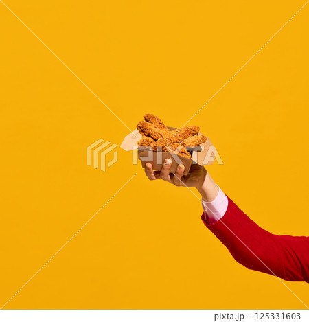 Raised hand holding crispy chicken nuggets box against yellow bold background. 125331603