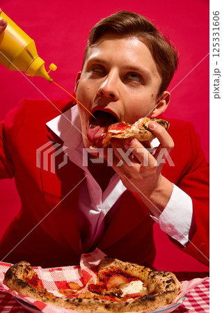 Man with delightful face pouring mustard while licking pizza slice against bold red background. 125331606