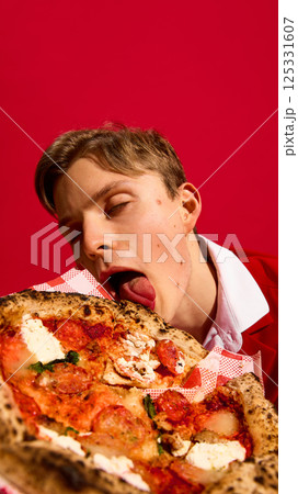 Young excited man with closed eyes licking pizza passionately against bold red background. 125331607
