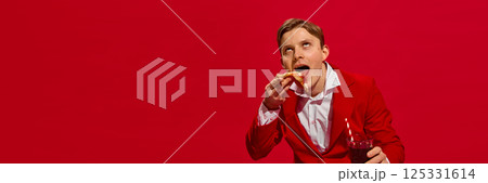 Man in red suit holding soda and dramatically biting pizza slice with ecstatic expression against bold red background. 125331614