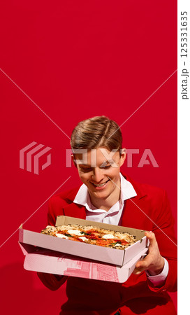 Smiling handsome man in red suit holding pizza box with excitement against vivid red background. 125331635
