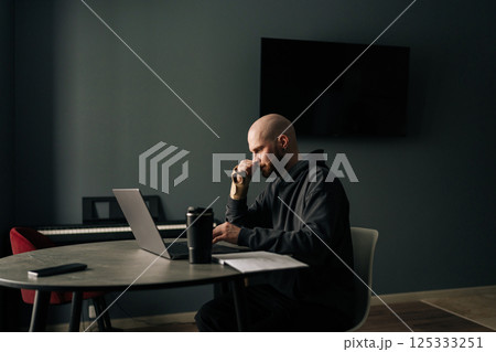 Focused freelancer with injured hand using laptop and drinking coffee while working from home office, sitting at desk with modern interior design, in cozy and productive workspace, looking to screen. 125333251