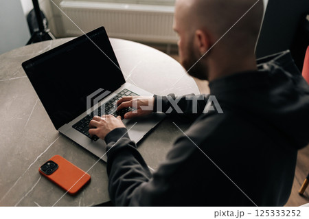View from shoulder of focused bald programmer with beard and hoodie sitting at table in home office, concentrating on work on laptop computer looking to screen, mobile phone nearby. View from shoulder of focused bald programmer with beard and hoodie sitting at table in home office, concentrating on work on laptop computer looking to screen, mobile phone nearby. 125333252