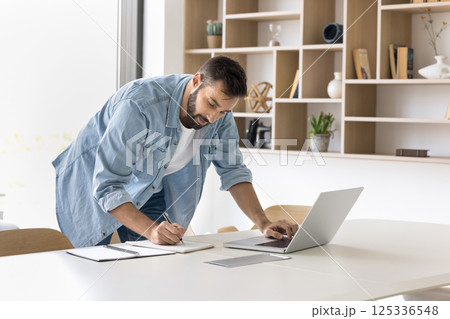 Focused millennial man stand by workplace browse internet write notes Focused millennial man stand by workplace browse internet write notes 125336548