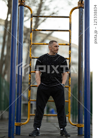 man wearing casual workout clothes stands confidently outdoor fitness park. He is positioned near exercise equipment surrounded urban scenery and greenery. man wearing casual workout clothes stands confidently outdoor fitness park. He is positioned near exercise equipment surrounded urban scenery and greenery. 125338834