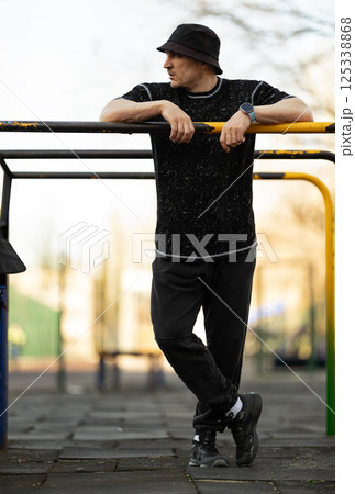 young man in black outfit and bucket hat stands casually against playground equipment. setting is park clear skies indicating vibrant day. young man in black outfit and bucket hat stands casually against playground equipment. setting is park clear skies indicating vibrant day. 125338868