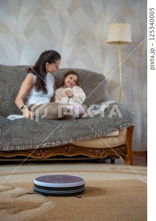 Robot vacuum cleaning the floor while a mother and daughter relax together on the couch in the background. Concept of smart home technology freeing time for family connection 125340005