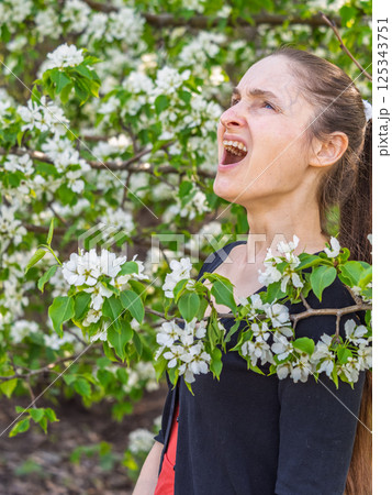 Young woman sneezes in the park against the background of a flowering tree. Allergy to pollen concept. 125343751