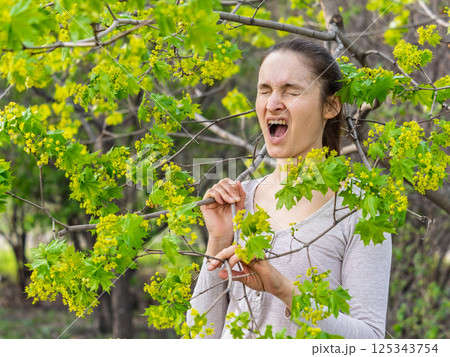 Young woman sneezes in the park against the background of a flowering tree. Allergy to pollen concept. Young woman sneezes in the park against the background of a flowering tree. Allergy to pollen concept. 125343754