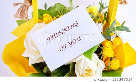 A vibrant basket filled with white and yellow flowers displays a card that reads Thinking of You. The setting suggests a warm and thoughtful gift, perfect for any occasion A vibrant basket filled with white and yellow flowers displays a card that reads Thinking of You. The setting suggests a warm and thoughtful gift, perfect for any occasion 125347514