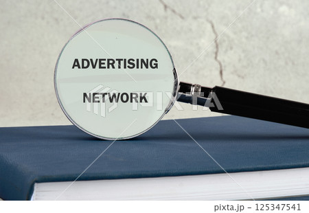 A magnifying glass highlighting the term advertising network placed on an open book against a textured wall background suggesting analysis and research 125347541