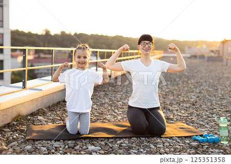 Young woman and girl showing biceps on rooftop after workout. Exercising together fosters strength, health, family bonding. Fitness equipment and urban nature background 125351358