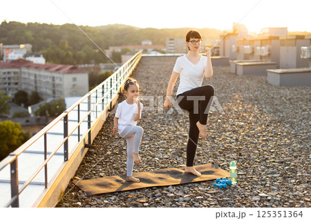 Young woman and little girl stretching during fitness workout on urban rooftop during sunset. Scene emphasizes health, bonding, outdoor activity and family connection. Young woman and little girl stretching during fitness workout on urban rooftop during sunset. Scene emphasizes health, bonding, outdoor activity and family connection. 125351364