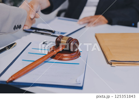 Two businesspeople are shaking hands in a law office with a scale and a gavel on the table 125351843