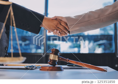 Two businesspeople are shaking hands in a law office with a scale and a gavel on the table 125351850