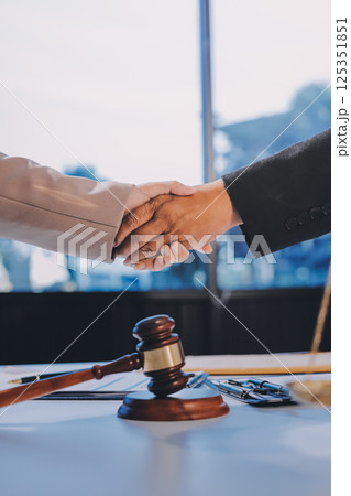 Two businesspeople are shaking hands in a law office with a scale and a gavel on the table 125351851