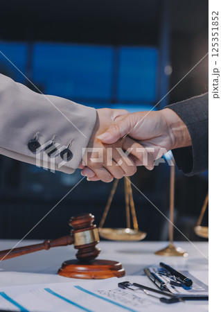 Two businesspeople are shaking hands in a law office with a scale and a gavel on the table 125351852