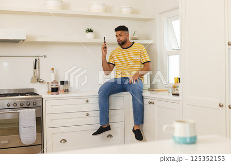 Young man in casual wear sitting on kitchen counter, using smartphone Young man in casual wear sitting on kitchen counter, using smartphone 125352153
