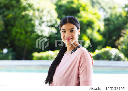 Smiling woman enjoying outdoor time, standing by pool with lush greenery 125352305