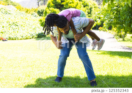 Father giving daughter piggyback ride in sunny park, both laughing together 125352343