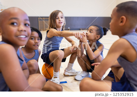 In school gym, diverse children sitting on basketball, talking and bonding as team In school gym, diverse children sitting on basketball, talking and bonding as team 125352915