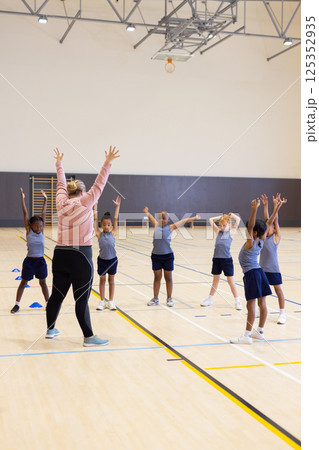 In school gym, female teacher leading group of diverse children in stretching exercises In school gym, female teacher leading group of diverse children in stretching exercises 125352935