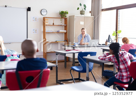 In school, female teacher sitting at desk, engaging with diverse students in classroom In school, female teacher sitting at desk, engaging with diverse students in classroom 125352977