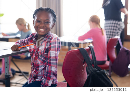 Smiling african american girl holding pencil in classroom with other students studying in school Smiling african american girl holding pencil in classroom with other students studying in school 125353011
